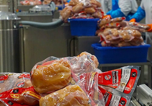 Packaged raw chicken sits in the foreground, with workers in the back working the line