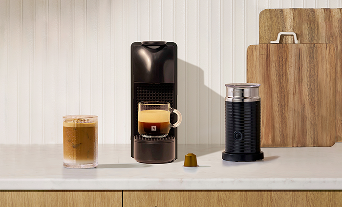 A black Nespresso Essenza Mini espresso machine with a milk frother, a single espresso in a glass mug, a gold coffee capsule, and an iced coffee, set against a background of light wood paneling and two large wooden cutting boards.