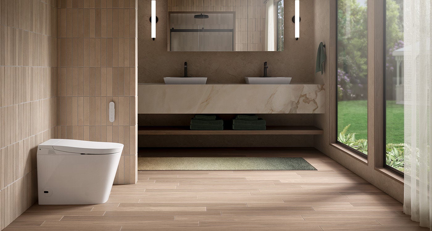 Nutural modern bathroom, showing a double sink vanity with white and tan marble counters. Left a mixed tan verticle tiled while with a white Kohler smart bidet leaned against the wall