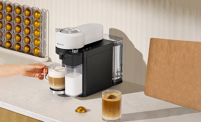 A white and black Nespresso Vertuo Lattissima machine brewing a latte into a glass mug, a pod holder filled with gold capsules, and a wooden cutting board in the background.