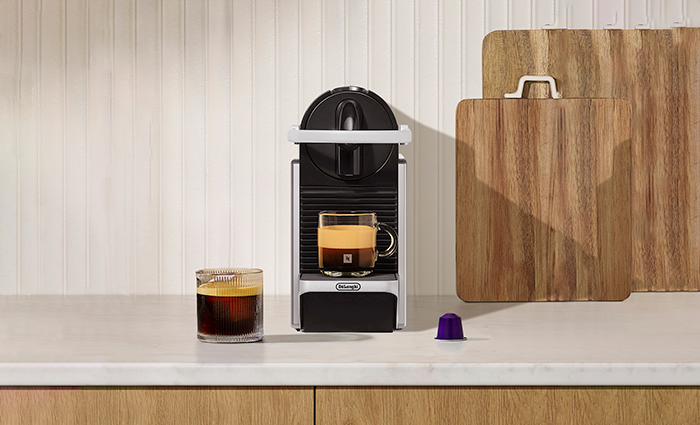 A silver Nespresso Pixie machine on a kitchen counter, featuring a brewed espresso in a glass mug, a purple coffee capsule, and wooden cutting boards in the background.