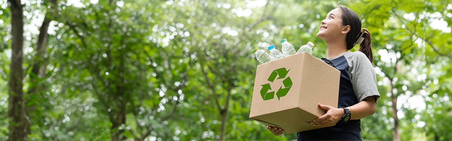 Young woman carrying recyclable box with circular recycling arrow through a green space outdoors