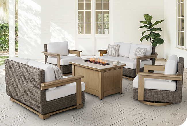 Modern wicker patio set with white cushions, wood accents, and a rectangular fire pit table.