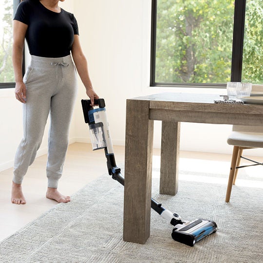Woman vacuuming with the cordless vacuum under a dining table.