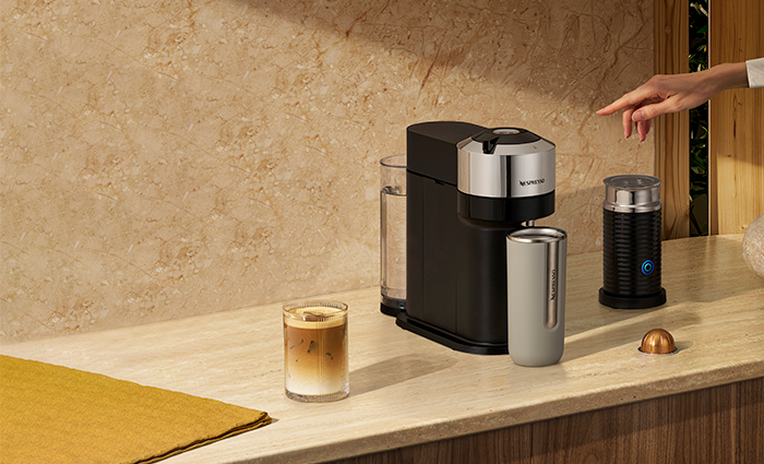 A Nespresso Vertuo Next coffee machine in a modern kitchen setting, accompanied by an milk frother, a travel mug, and a glass of iced coffee.